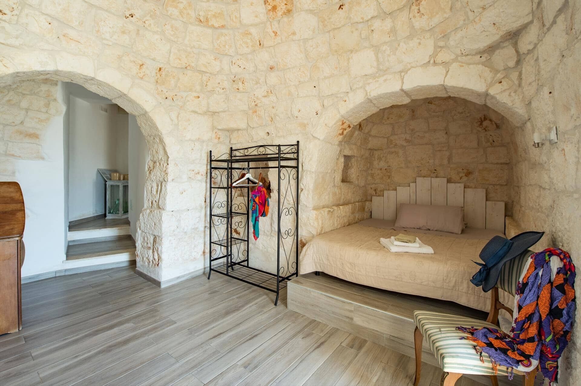 A bedroom with a stone wall, a bed with a headboard, a metal coat rack, and a wooden bench.
