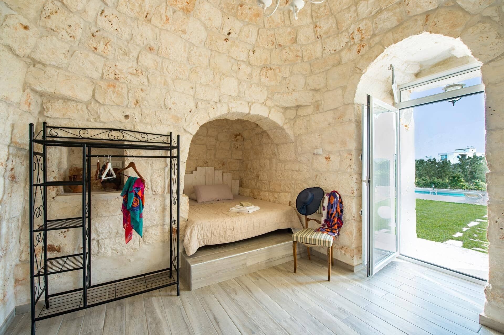 A room with a stone wall, a bed, a metal rack with clothes, a chair, and a view of a pool through a glass door.