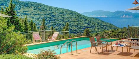 A pool with a view of the sea and mountains, surrounded by outdoor furniture and greenery.