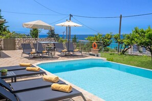 Ein Schwimmbecken mit Liegestühlen und einem Tisch, umgeben von einer Steinmauer und Blick aufs Meer.