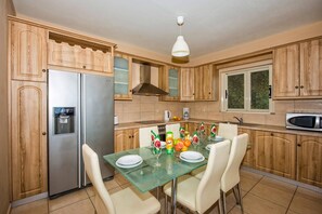 A kitchen with wooden cabinets, a stainless steel refrigerator, a glass dining table with chairs, and a hanging light fixture.