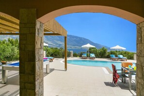 Ein Poolbereich mit Liegestühlen und Sonnenschirmen, ein steinernes Tor und Blick auf das Meer und die Berge.