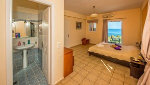 A bedroom with a bed, a bedside table, a nightstand, a window with a view of the sea, and a bathroom with a sink and mirror.