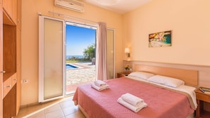 A hotel room with a bed, a nightstand, a lamp, a painting, and a view of a pool and garden. - Villa Skala Ionian by Villa Plus (Ionian Islands)