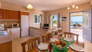 A well-lit kitchen with modern appliances, a dining area with a table set for two, and a view of the sea through the open door. - Villa Skala Ionian by Villa Plus (Ionian Islands)