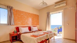 Um quarto com uma cama grande, uma mesa de cabeceira, uma janela com cortinas e vista para o mar através de uma porta aberta.
