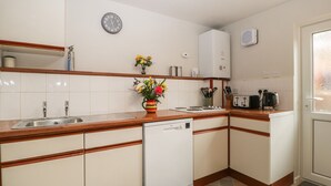 A compact kitchen with white cabinets, a built-in dishwasher, a stove, and a sink. There is a clock on the wall and a small vase with flowers on the counter.