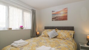 A neatly made bed with floral bedding, a window with curtains, a wall-mounted painting, and a bedside table with a lamp.