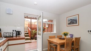 A compact kitchen with a toaster, kettle, and a small dining table with chairs. A window with a view of a garden and a door leading outside.