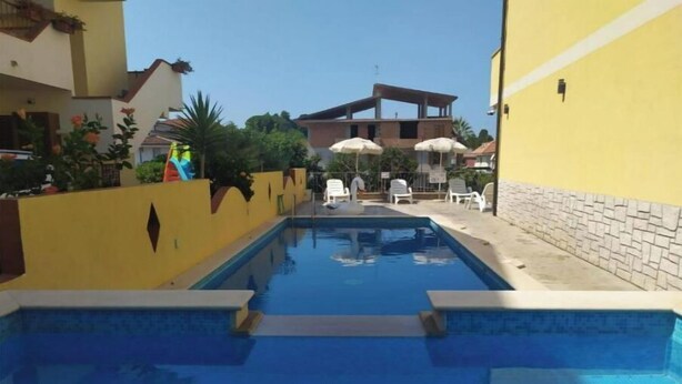 A swimming pool with a clear blue water surface, surrounded by a yellow wall and a stone wall. There are white lounge chairs and umbrellas in the background. - PLAYA DEL SOL Three-room apartment 1 (Santa Domenica)