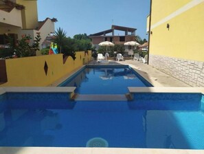 Une piscine à l’eau d’un bleu limpide, entourée d’un mur jaune et d’un mur en pierre. On aperçoit, au fond, des chaises longues blanches et des parasols.