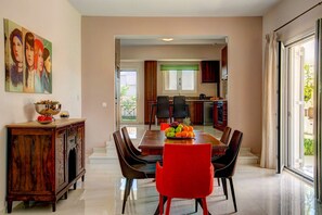Un coin repas avec une table en bois, des chaises rouges et un saladier de fruits. On aperçoit une cuisine au fond, avec une fenêtre et un balcon.