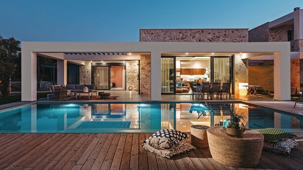 Una casa moderna con piscina, mobiliario de exterior y cocina visible a través de puertas de cristal.