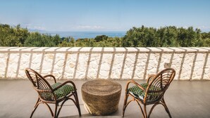A balcony with two wicker chairs and a round wicker ottoman, overlooking a coastal landscape with trees and the sea. - Villa Tiare 2 by Villa Plus (Ionian Islands)