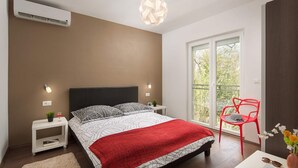 Une chambre à coucher avec un lit, des tables de chevet, un fauteuil rouge, une fenêtre et un appareil de climatisation.