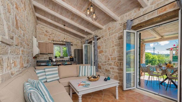 A cozy living room with a stone wall, a sofa with striped pillows, a coffee table with magazines, and a view of an outdoor patio with a table and chairs. - Villa Rizolia Enea by Villa Plus (Koridallos)
