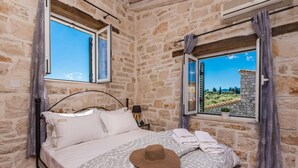 A stone-walled bedroom with a bed, a window with a view of greenery, and a hat on the bed. - Villa Rizolia Enea by Villa Plus (Koridallos)