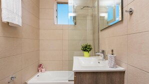A modern bathroom with a bathtub, a sink, and a shower. There is a potted plant on the sink counter and toiletries on the side. - Villa Rizolia Enea by Villa Plus (Koridallos)