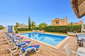Piscina com espreguiçadeiras, um telhado de palha e um edifício com telhado de azulejos.