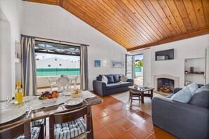A living room with a wooden ceiling, a fireplace, a sofa, and a dining area with a table set for two. - Villa Sunshine by Villa Plus (Albufeira)