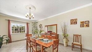 A dining area with a wooden table and chairs, a chandelier, a window with curtains, and wall-mounted artwork. - Villa Solange by Villa Plus (Albufeira)