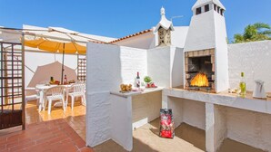 En udendørs terrasse med røde mursten, hvide vægge, et hvidt bord med stole, en gul parasol, en murstensovn og en pose kul.
