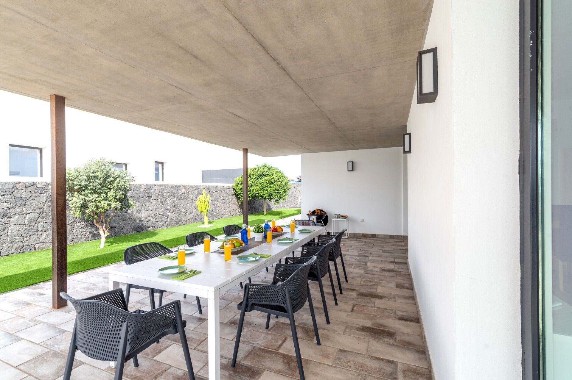 A covered outdoor dining area with a long white table, black chairs, and a stone wall in the background.