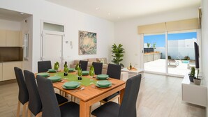 Un espace de repas moderne avec une table en bois dressée pour quatre personnes, entourée de chaises sombres. Une grande fenêtre offre une vue sur une piscine et un ciel dégagé.