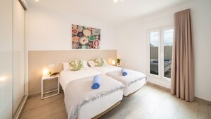 Une chambre d’hôtel avec deux lits, une grande fenêtre avec des rideaux, une œuvre d’art accrochée au mur et une table de chevet avec une lampe.