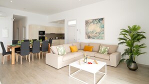 Un salon moderne avec un canapé beige, un coin repas avec une table et des chaises en bois, une cuisine en arrière-plan et une grande plante en pot.