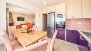 Een moderne keuken met een inbouwoven, een koelkast en paarse kastjes. Er is een eethoek met een houten tafel en roze stoelen.