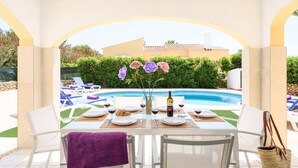 A poolside dining area with a table set for two, a bottle of wine, and a vase with flowers. - Villa Sol I by Villa Plus (Cap d'Artrutx)