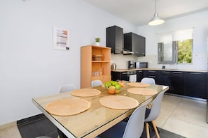 A modern kitchen with black cabinets, a wooden dining table with woven placemats, and a view of greenery outside. - Villa Rhodes Sea View II by Villa Plus (Ixia)