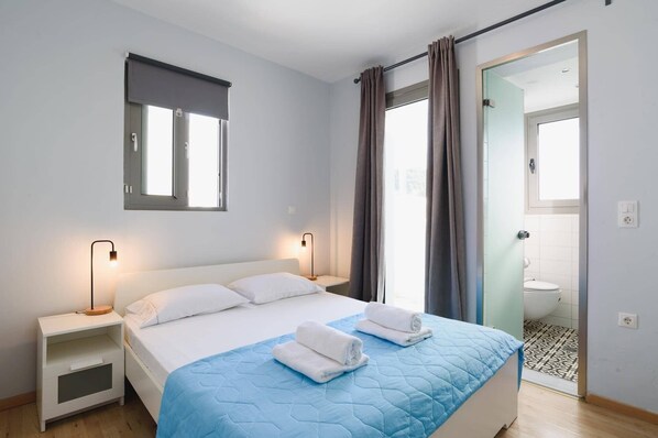 A bedroom with a bed, bedside tables, a window with curtains, and a bathroom visible through an open door.