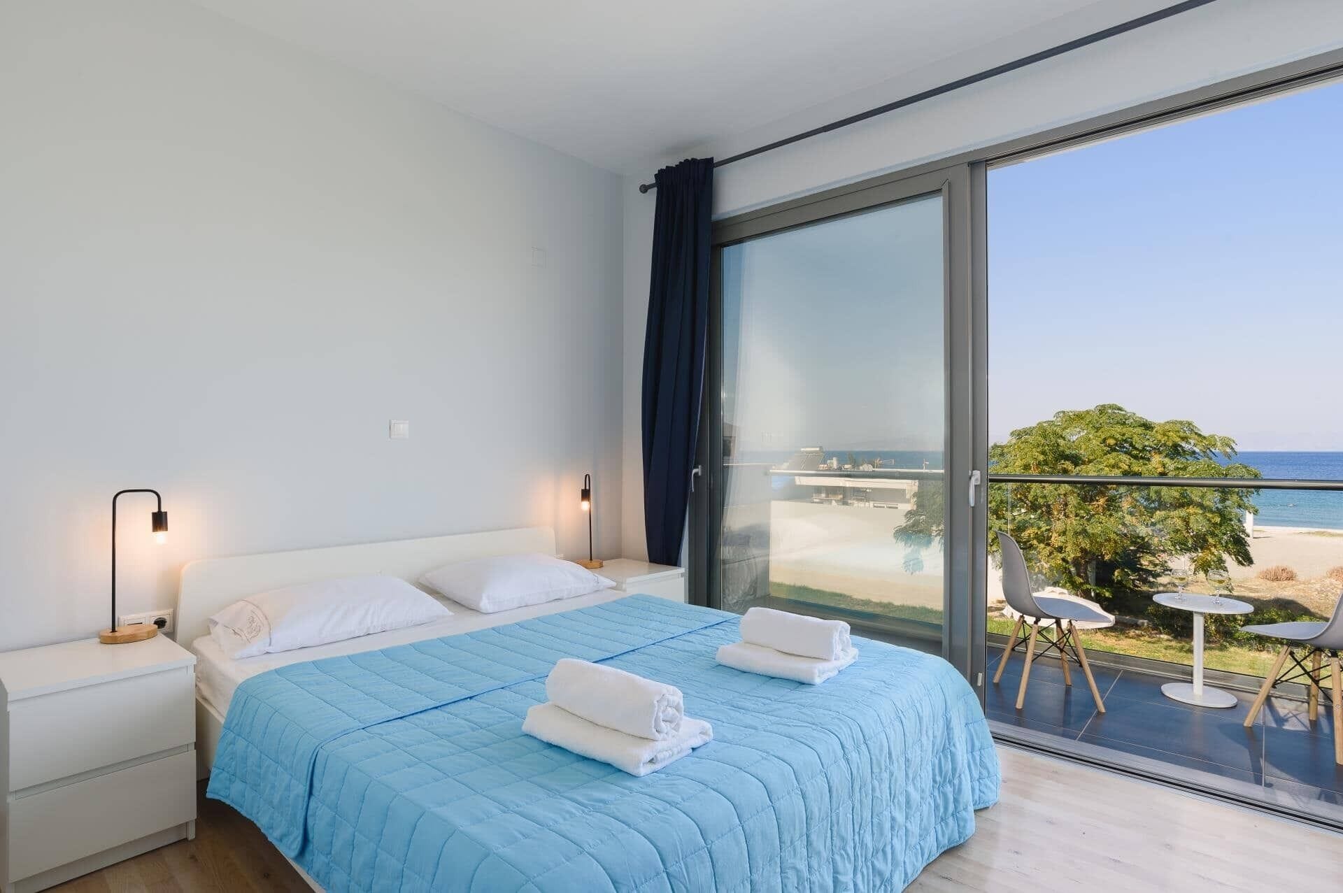 Quarto de hotel moderno com uma cama grande, duas luminárias de cabeceira, vista para a praia e uma varanda com área de estar externa.