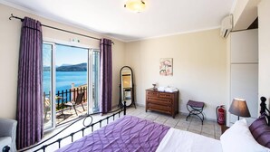 Une chambre d’hôtel avec un lit, une commode, un miroir, une chaise, une petite table, une lampe et une vue sur la mer.
