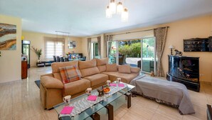 A spacious living room with a beige sofa, a glass coffee table, and a television. There is a dining area in the background with a table and chairs. - Villa Paraisos Sol by Villa Plus (Las Lagunas de Mijas)