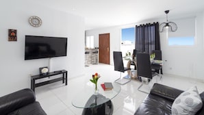 A modern living room with a flat-screen TV, a glass coffee table with flowers, a leather sofa, and a dining area with a glass table and chairs. - Villa Sunny Side by Villa Plus (Puerto del Carmen)