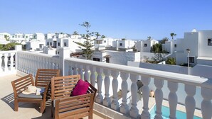 A balcony with wooden chairs and a white balustrade overlooking a coastal residential area with white buildings and a clear blue sky. - Villa Sunny Side by Villa Plus (Puerto del Carmen)