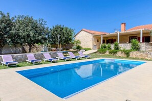 Une piscine entourée de chaises longues, un bâtiment en pierre avec un toit en tuiles et une végétation luxuriante.
