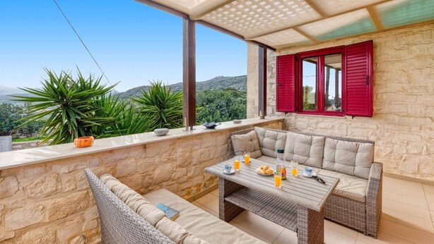 Ein Balkon mit einer Rattan-Couch, einem Tisch mit Getränken und Blick auf Grünflächen und Hügel.