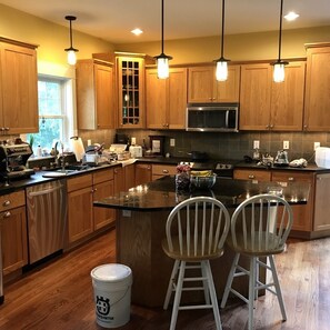 A modern kitchen with wooden cabinets, a central island with two chairs, a microwave, and various kitchen appliances. - Solar-eV Country Home - Quaint Cazenovia! (Cazenovia)