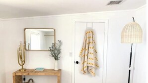 A hallway with a wooden console table, a mirror, a vase with greenery, a hanging towel, a floor lamp, and a woven basket. - Joshua Tree - Under the Stars-Pool, Spa & Fire pit (Yucca Valley)