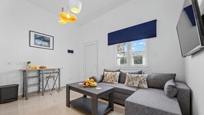 A modern living room with a grey sofa, a small table with fruit, a chair, a TV, and a window with a view of the outside. - Home2Book Charming Apartment Teguise, Terrace (Nazaret)