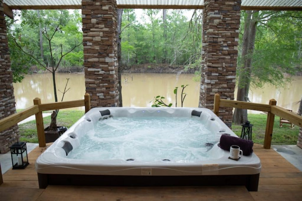 Un bain à remous doté d’une terrasse en bois et de piliers en pierre, entouré d’une clôture en bois, et offrant une vue sur une rivière.