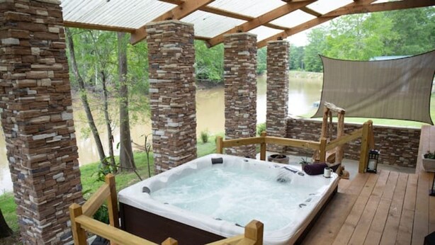 A hot tub with a wooden deck and stone pillars, surrounded by a covered area with a view of a lake.
