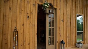 A wooden building with a door open to a room, a lantern on the porch, and a small shelf with items.