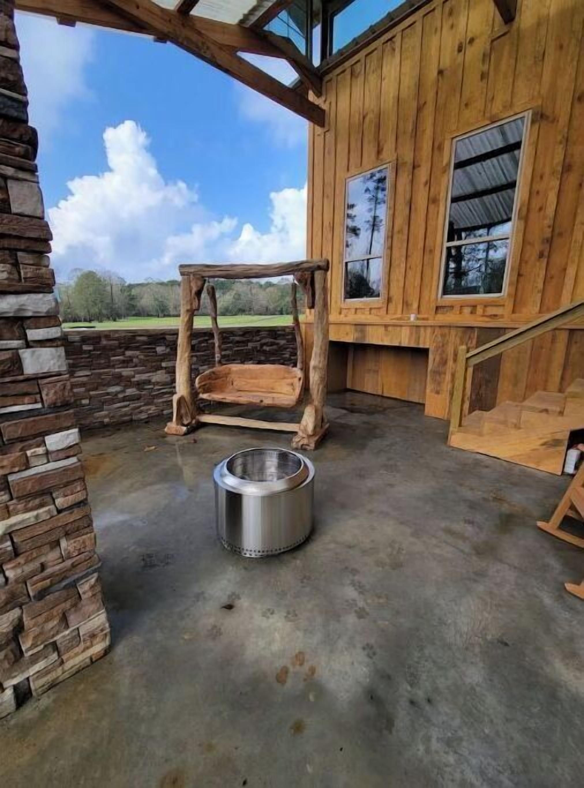 A rustic wooden cabin with a stone wall, a metal bowl on the floor, and a wooden swing.