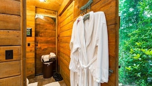 Une salle de bain rustique, avec des murs et une porte en bois, un porte-serviettes sur lequel sont disposés des peignoirs blancs, et une petite poubelle.