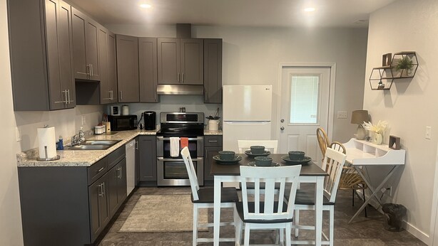 Una cuina moderna amb armaris foscos, electrodomèstics d'acer inoxidable i zona de dinar amb cadires blanques i una taula.